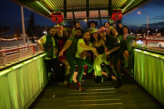 Group of friends in cowboy hats and bandanas partying on a green-lit open-air pedal pub on a city bridge at night, holding drinks and posing for a photo