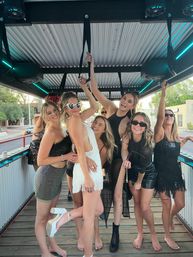 Group of women in black and white party dresses and sunglasses posing and laughing on a covered outdoor party trolley deck with hand straps, neon accent lights and a city street with trees visible outside.