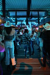 Nighttime neon-blue country bar scene with people in cowboy hats and boots dancing on a wooden floor under a corrugated metal roof