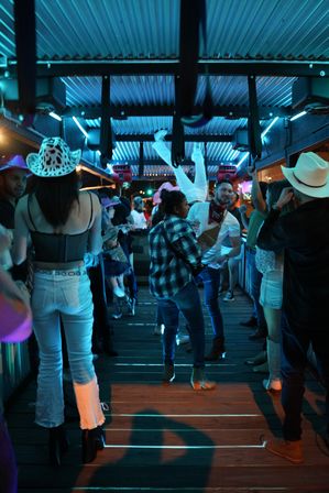 Nighttime neon-blue country bar scene with people in cowboy hats and boots dancing on a wooden floor under a corrugated metal roof