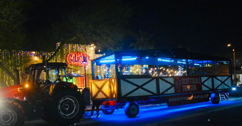 Nighttime tractor-pulled party wagon glowing with blue LED underglow and string lights, festive riders and decorated storefronts along a street.
