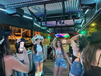 Friends dancing on a lively outdoor rooftop patio at night, wearing denim shorts and cowboy hats under colorful LED lights with city street traffic in the background.