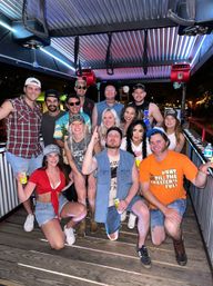 Smiling group of friends celebrating on a covered outdoor party pedal bar at night, wearing casual summer outfits and holding drinks.