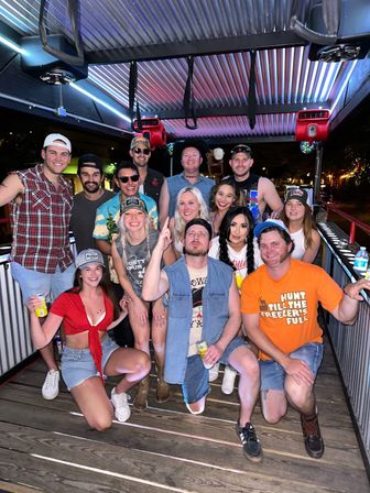 Smiling group of friends celebrating on a covered outdoor party pedal bar at night, wearing casual summer outfits and holding drinks.