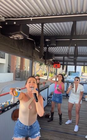 Four women having a daytime city-side party on a covered outdoor trolley — one sings into a microphone while holding a canned drink, others laugh, film on a phone and hold drinks, wearing casual summer outfits and sunglasses.