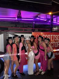 Seven women in pink outfits and cowboy hats, one wearing white sash and hat, smiling and posing at night under purple neon lights beside a decorated party trailer.