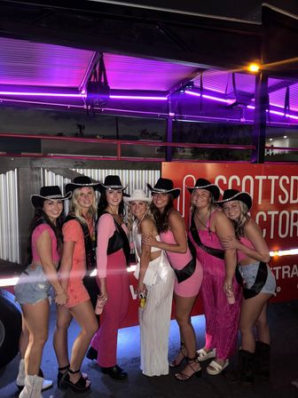 Seven women in pink outfits and cowboy hats, one wearing white sash and hat, smiling and posing at night under purple neon lights beside a decorated party trailer.