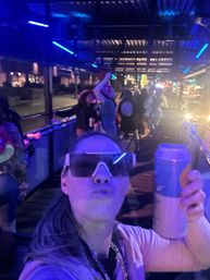 Neon-lit city nightlife selfie on an outdoor patio — person in oversized visor holding a canned drink, crowded covered walkway and street party in the background.