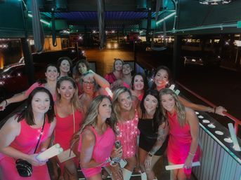 Smiling group of women in pink outfits on an outdoor rooftop bar patio at night, holding glowing LED light sticks and posing under neon lights for a lively girls' night out.