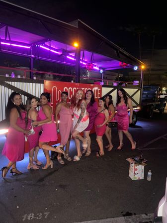 Group of women in pink dresses posing and smiling at night in front of a neon-lit outdoor mobile bar trailer in Scottsdale, Arizona — festive bachelorette-style celebration.