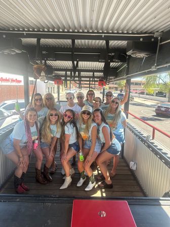 Group of a dozen women in matching tees, denim shorts and cowboy boots smiling and posing with drinks on an open-air party trolley under a corrugated metal roof in a sunny urban parking area.