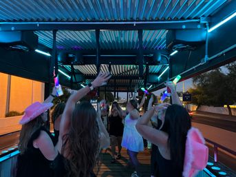 Friends dancing on an illuminated outdoor party bike at dusk with neon blue LED lights, pink cowboy hats and glowing bottles, city street in the background