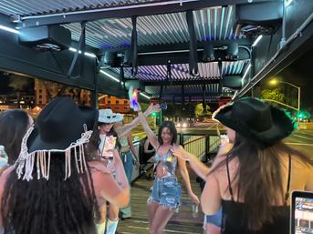 Group of young women dancing on a covered outdoor patio at night, wearing cowgirl hats and festival outfits under colorful LED lights with a downtown street in the background.