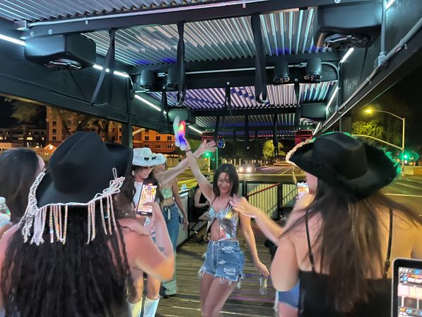 Group of young women dancing on a covered outdoor patio at night, wearing cowgirl hats and festival outfits under colorful LED lights with a downtown street in the background.
