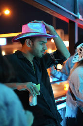 Neon-lit urban nightlife scene: a man wearing a glittery pink cowboy hat leans at a rooftop bar at night, holding a canned drink under city lights.