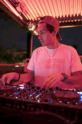 Rooftop DJ mixing on a controller under pink-red neon lights, wearing a backward cap and cactus graphic tee — lively nighttime rooftop scene.