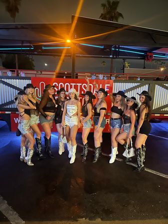 Group of women in cowgirl hats, denim shorts and boots posing at night in front of a Scottsdale sign with neon lights