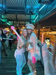 Two friends smiling on a neon-lit outdoor patio at night, one wearing a sparkly hat and holding a toy hobby horse and pink inflatables, with a phone photographer and city lights in the background for a lively nightlife celebration.