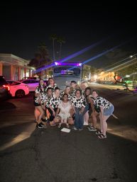 Group of women in matching cow-print shirts and one in white lace posing at night on a palm-lined downtown street in front of a lit-up truck with purple party lights
