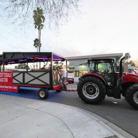 Red tractor towing an open-air party wagon lit with purple LED lights as passengers board on a palm-lined suburban street at dusk.