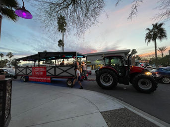 Red tractor pulling an open-air party wagon down a palm-lined southwestern desert city street at sunset, neon lights glowing and people boarding.