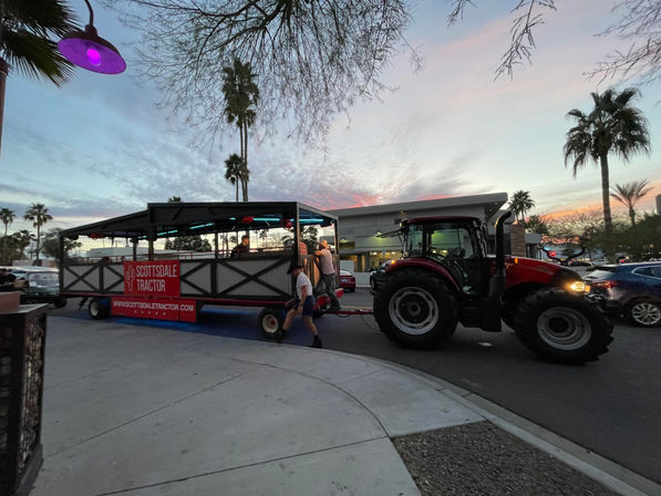 Red tractor pulling an open-air party wagon down a palm-lined southwestern desert city street at sunset, neon lights glowing and people boarding.