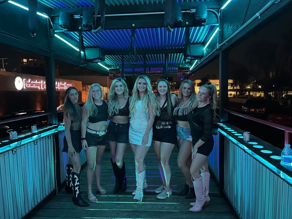 Seven women posing on an illuminated outdoor rooftop bar deck in city nightlife, under teal neon lights, wearing party outfits and cowboy boots.