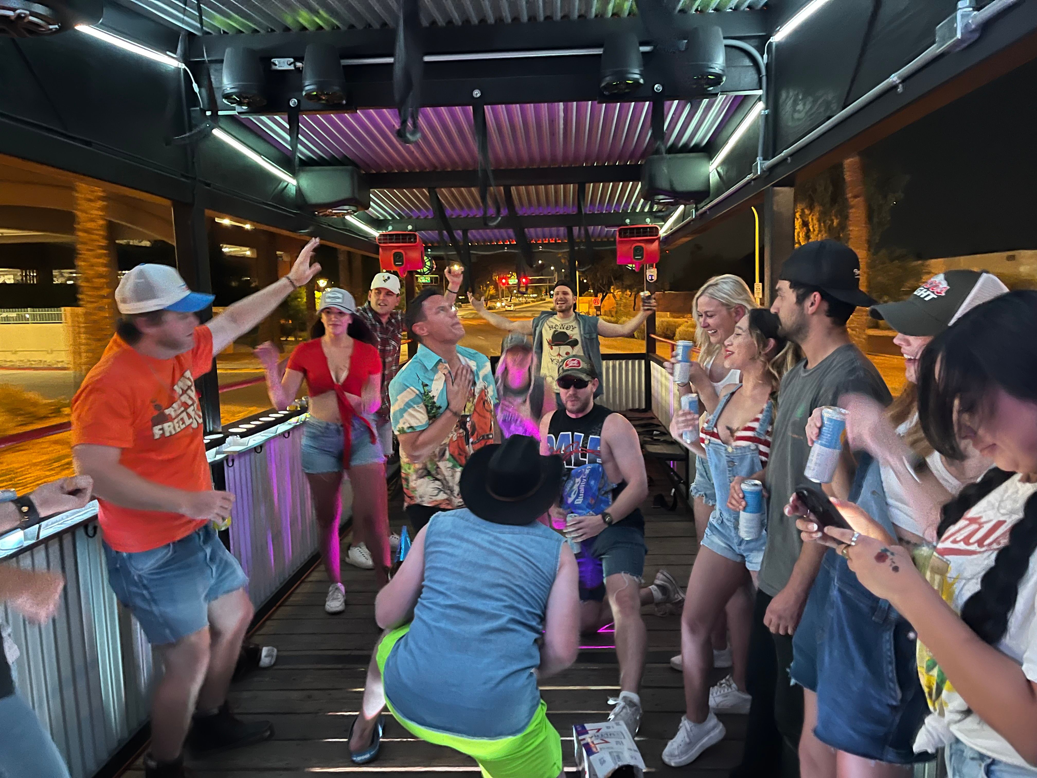 Group of people dancing and laughing on a covered outdoor patio at night with colorful LED lights, casual summer outfits, drinks in hand and a city street visible beyond.