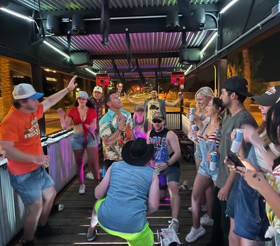 Group of people dancing and laughing on a covered outdoor patio at night with colorful LED lights, casual summer outfits, drinks in hand and a city street visible beyond.