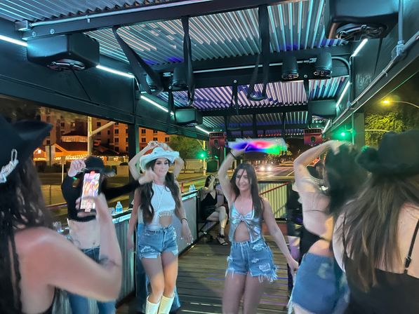 Group of women dancing on a covered outdoor patio at night with neon strip lights and speakers overhead, wearing cowboy hats and denim shorts, downtown street visible