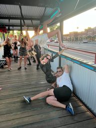Group of friends partying on an open-sided trolley at sunset on a city street; a woman in a white fringe outfit hangs upside down from overhead straps while others cheer and a man in sunglasses lounges on the wooden deck pointing at the camera.