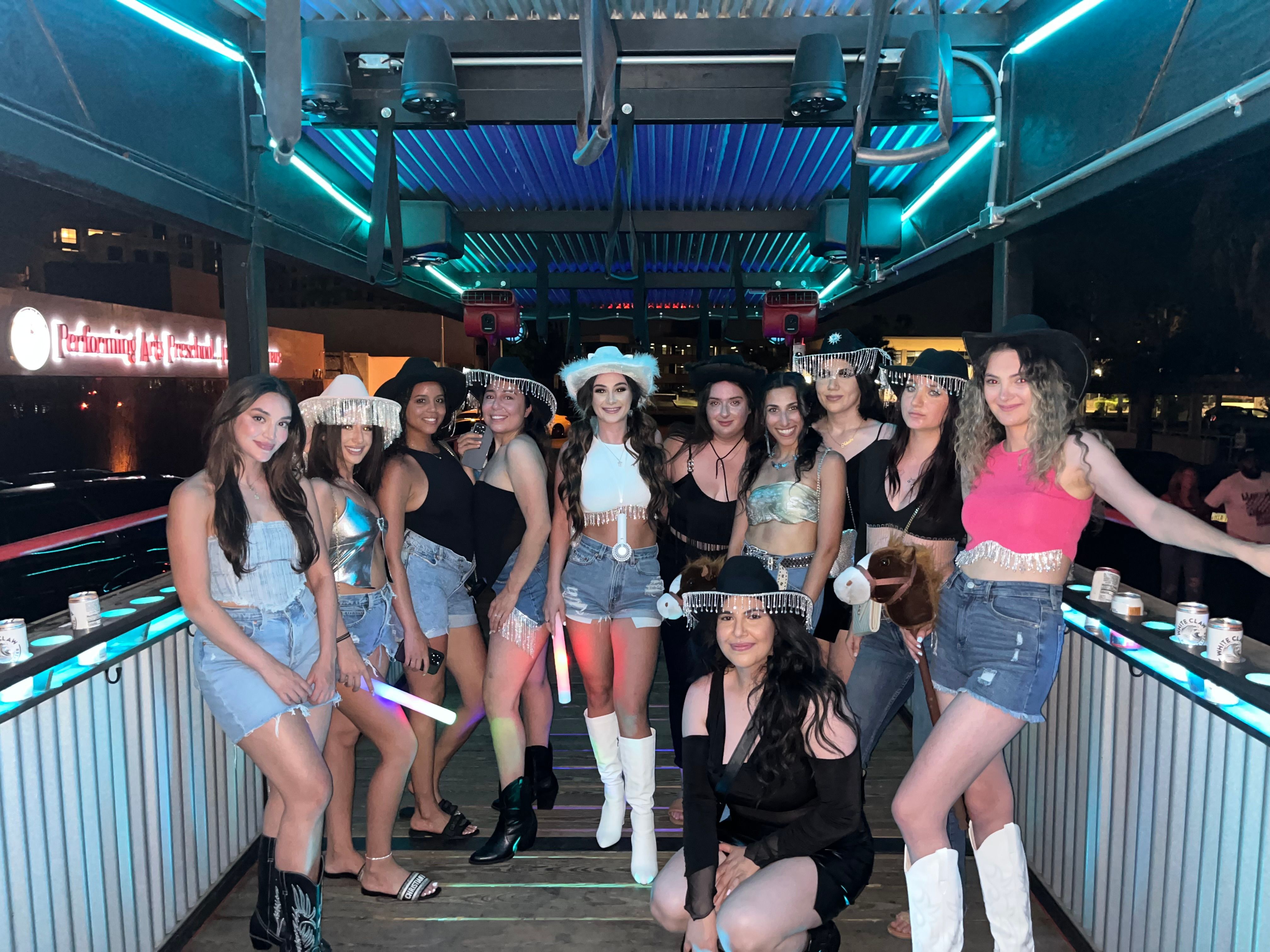 Group of friends in cowgirl outfits and denim posing with cowboy hats, fringe, hobby horses, and glow sticks on a neon-lit outdoor bar patio at night.