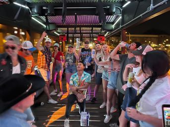 Group of friends partying on a covered outdoor bar deck at night, holding drinks, dancing and cheering under neon lights with downtown street lights in the background.