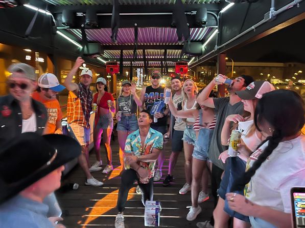 Group of friends partying on a covered outdoor bar deck at night, holding drinks, dancing and cheering under neon lights with downtown street lights in the background.