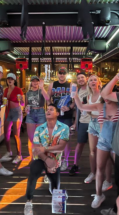 Group of friends partying on a covered outdoor bar deck at night, holding drinks, dancing and cheering under neon lights with downtown street lights in the background.