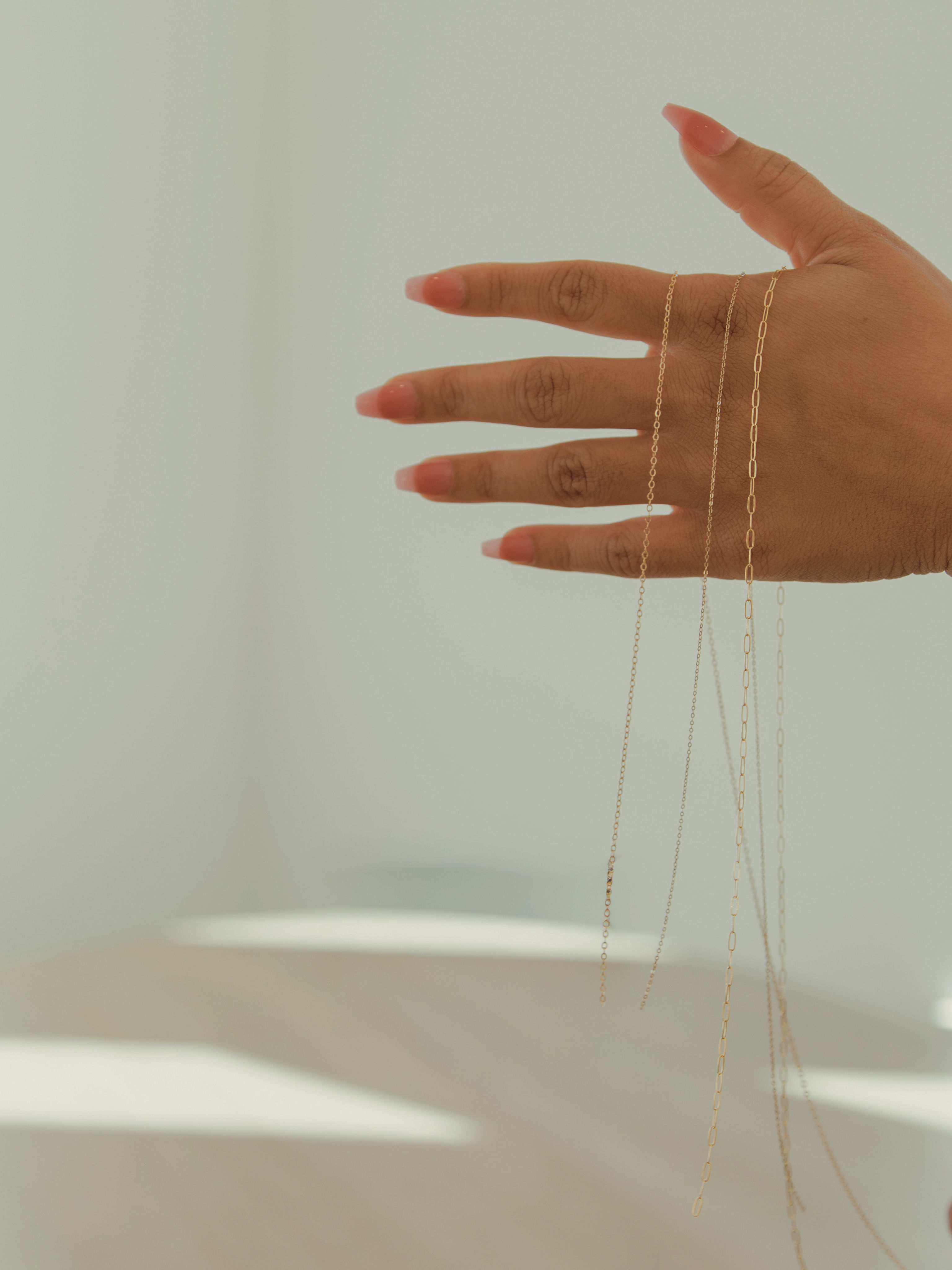 Manicured hand dangling thin gold necklace chains against a soft, sunlit neutral background.