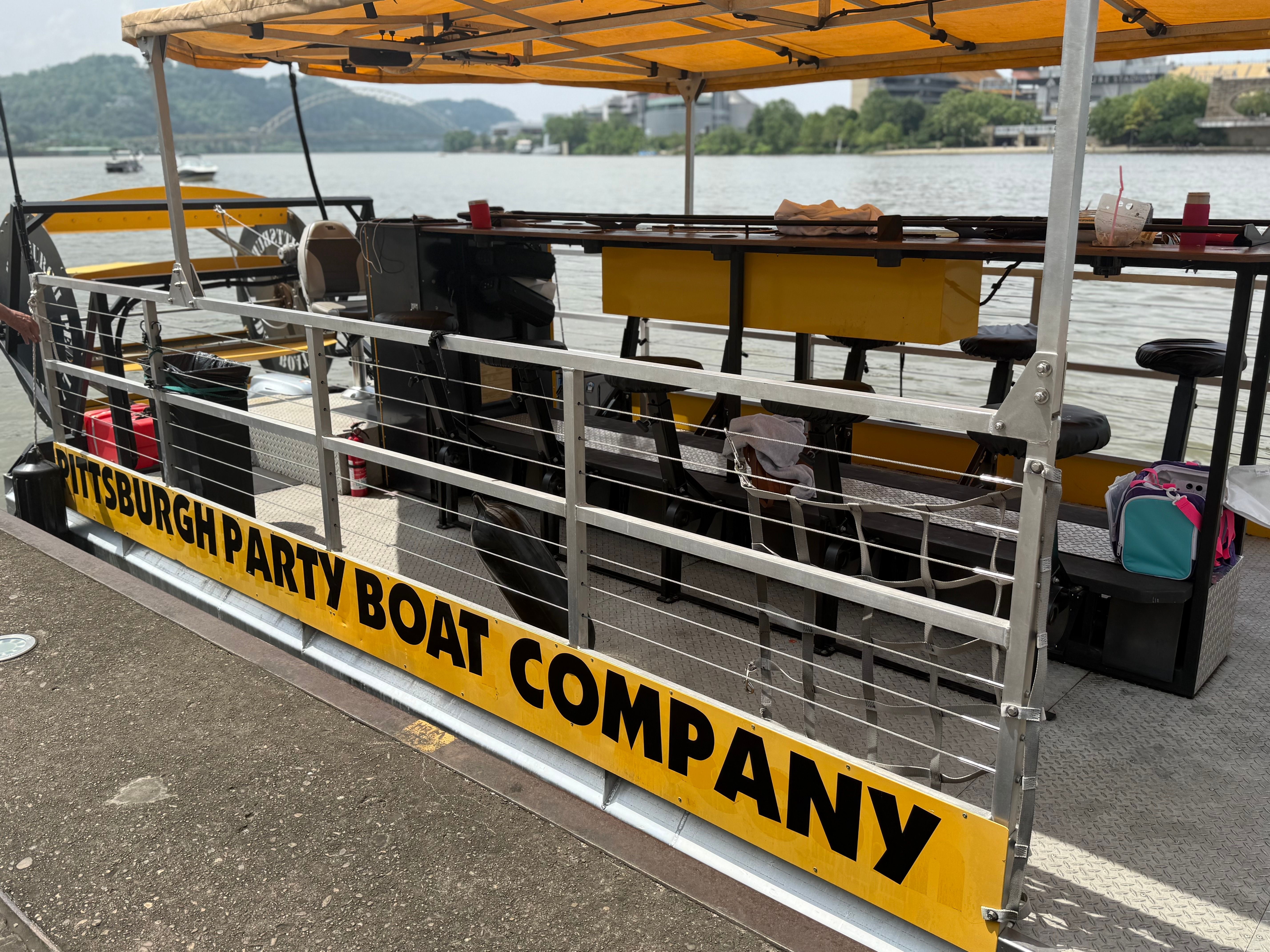Yellow-canopied party boat docked on the Pittsburgh riverfront with metal railings, bar stools on deck, and a bridge and green hills across the river