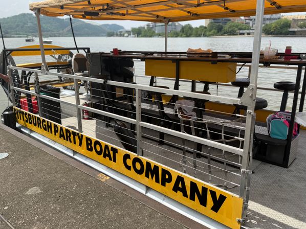 Yellow-canopied party boat docked on the Pittsburgh riverfront with metal railings, bar stools on deck, and a bridge and green hills across the river