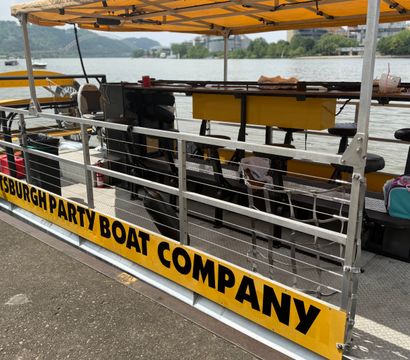 Yellow-canopied party boat docked on the Pittsburgh riverfront with metal railings, bar stools on deck, and a bridge and green hills across the river