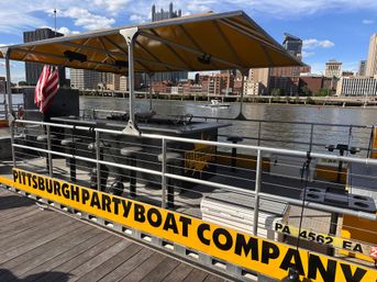 Yellow-canopied party pontoon docked on the Pittsburgh riverfront with a bar counter, stools, American flag and city skyline across the river.