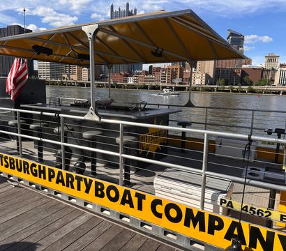 Yellow-canopied party pontoon docked on the Pittsburgh riverfront with a bar counter, stools, American flag and city skyline across the river.