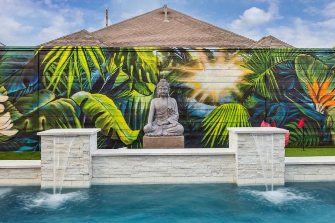 Zen Buddha statue centers a backyard scene: vibrant tropical mural on a fence, twin stone water spouts cascading into a blue swimming pool, with house roofs in the background.