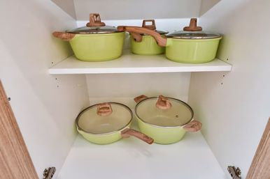 Five-piece light green cookware set with wood-look handles and glass lids neatly arranged on two shelves inside a white kitchen cabinet
