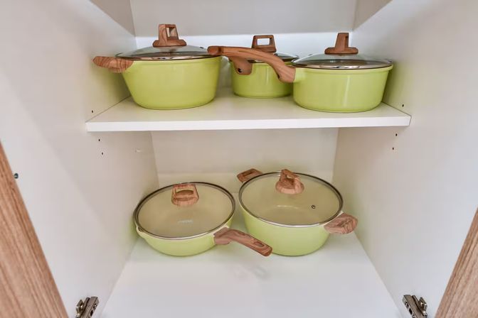 Five-piece light green cookware set with wood-look handles and glass lids neatly arranged on two shelves inside a white kitchen cabinet