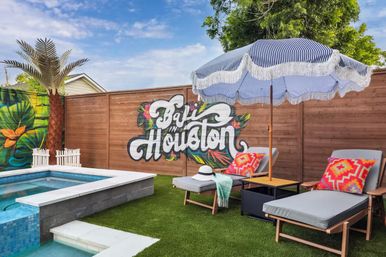 Houston backyard pool lounge with tropical mural reading "Bali in Houston", blue-striped fringe umbrella, two chaise lounges with colorful pillows, small pool, artificial turf and palm accent