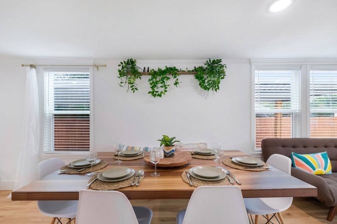 Sunlit modern dining room with a wooden table set for six, white chairs, woven placemats, trailing shelf plants and large windows