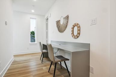 Bright modern home office nook with a built-in gray double desk, two chairs, light wood floors, tall slim window, and boho rattan sunburst and oval mirrors on a white wall.