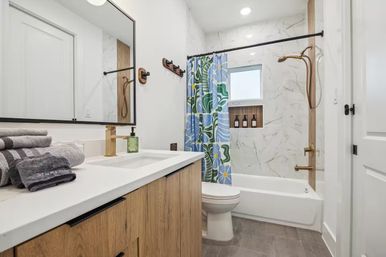 Contemporary residential bathroom with wood vanity and white countertop, stacked gray towels, large framed mirror, brass faucet and shower fixtures, marble-look tub surround, blue-green floral shower curtain, recessed niche with bottles and gray tile floor.