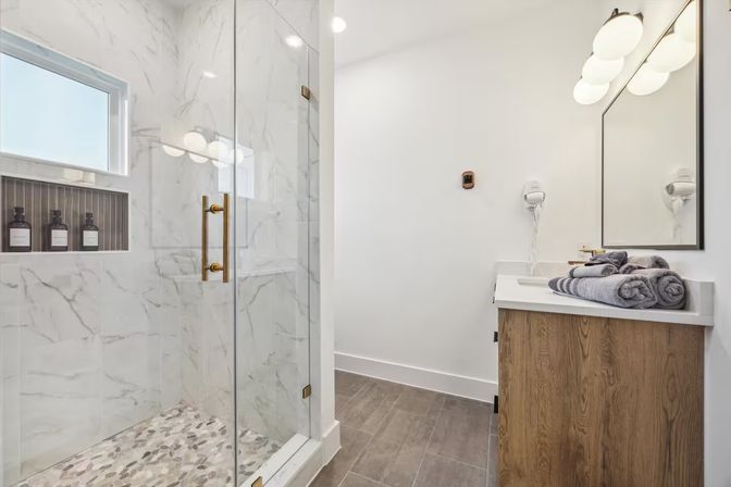 Bright modern bathroom interior with glass-enclosed marble shower and pebble floor, brass hardware, recessed shampoo niche, wood vanity with white countertop, wall mirror and neatly rolled gray towels.
