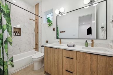 Bright modern bathroom featuring marble-look shower tile, brass rain shower, tropical green leaf shower curtain, white tub and toilet, wood double-sink vanity with quartz countertop, large rectangular mirror and hanging plant.